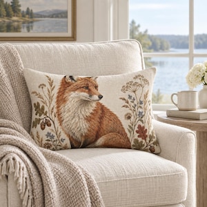 May include: A rectangular throw cushion featuring a woven illustration of a red fox centred among botanical elements including oak leaves, acorns, and wildflowers. The design is rendered in natural earth tones against a light beige background.