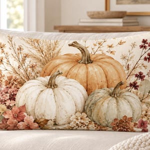 May include: A rectangular throw pillow featuring a printed illustration of three pumpkins in shades of orange, white, and muted green. The design includes dried grasses, autumn leaves, and small floral accents in warm brown and rust tones.