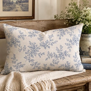 Puede incluir: Un cojín rectangular con una base de tela de color beige claro y un patrón repetitivo de helechos en azul. El diseño consiste en hojas delicadas y ramificadas extendidas por toda la superficie, creando un motivo floral consistente.
