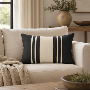 May include: A rectangular throw pillow featuring a wide central cream-colored vertical stripe flanked by two thinner black stripes on each side, set against a solid black background. The pillow has a smooth, matte fabric texture and clean edges.