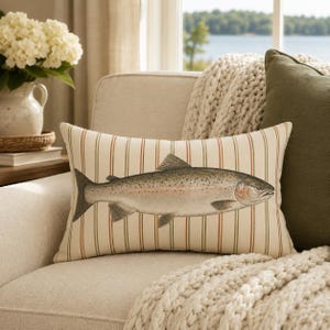 May include: A rectangular throw pillow featuring a cream-colored fabric with vertical stripes in thin red and green lines. Centered on the pillow is a detailed illustration of a fish with silver, grey, and pinkish scales and dark spots.