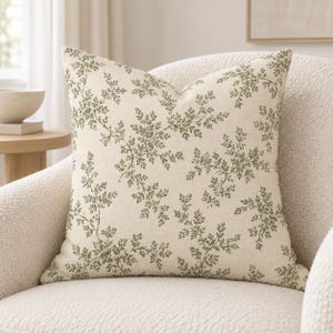 May include: A square throw pillow featuring a cream-colored fabric base with a repeating dark green botanical fern leaf pattern. The pillow has a textured, woven appearance and is displayed centered on a textured off-white upholstered chair.
