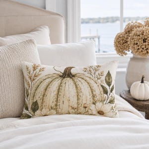 Peut inclure: Un coussin rectangulaire orné d'une illustration imprimée d'une grande citrouille vert pâle entourée d'épis de blé, de feuilles vertes et de fleurs blanches. Le coussin présente un fond couleur crème et est posé sur un lit avec d'autres coussins.