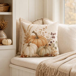 May include: A square throw cushion featuring a printed illustration of three pumpkins in shades of orange, white, and muted green, surrounded by dried wheat stalks, autumn foliage, and small branches on a cream-coloured fabric background.
