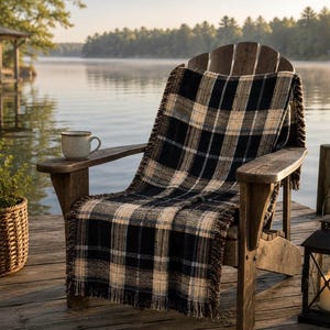 May include: A woven throw blanket featuring a black, cream, and tan plaid pattern with fringed edges, draped over a wooden Adirondack chair. The blanket has a textured appearance and covers the seat and backrest of the chair in an outdoor setting.