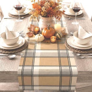 May include: A table runner featuring a tan, white, and black plaid pattern centered on a wooden table. The table is set with white bowls, woven placemats, wine glasses, and a centerpiece of autumn foliage, pumpkins, and lit candles in glass holders.