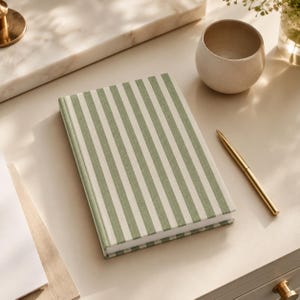 May include: A rectangular notebook featuring a vertical striped pattern in sage green and off-white. The cover has a fabric-like texture. A slim gold-toned metal pen is positioned diagonally next to the notebook on a smooth, light-colored surface.