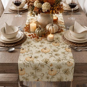 May include: A cream-colored table runner featuring a repeating pattern of orange pumpkins and green vine leaves. The runner is displayed on a wooden dining table set with ceramic bowls, plates, wine glasses, and a centerpiece of autumn foliage.