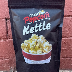 May include: Black bag of Pride Popcorn Kettle popcorn. The bag features the words "Pride Popcorn Kettle" in red and white text. The bag shows a red and white striped container filled with yellow popcorn.