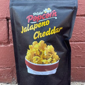 May include: A black bag of Pride Popcorn Jalapeno Cheddar. The bag features the brand name in red and white, with the flavor in yellow. A bowl of yellow popcorn with red and white trim is pictured.