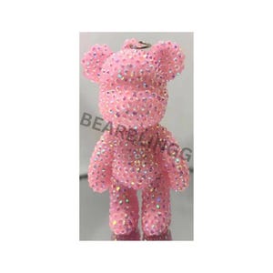 Könnte beinhalten: Rosa, mit Strass besetztes Bärenfigur mit einer silbernen Schlaufe oben. Der Bär ist mit schillernden Strasssteinen bedeckt, und der Text "BEARBLINGG" ist auf der linken Seite sichtbar. Der Bär hat eine klassische Teddybärform.