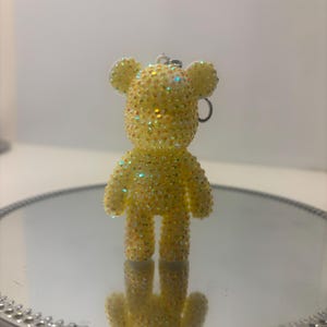 Op de afbeelding: Een gele, met glitter bedekte sleutelhanger in de vorm van een beer. De beer heeft een ronde kop met kleine oren en een sleutelring. Het oppervlak is bedekt met kleine, sprankelende versieringen. De sleutelhanger wordt weerspiegeld in een ronde spiegel.