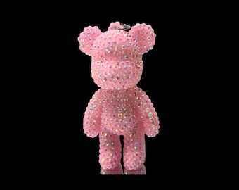 Pode incluir: Uma estatueta em forma de urso coberta com pequenos strass cor-de-rosa claro iridescentes que refletem a luz. O urso tem orelhas arredondadas, um focinho definido e um pequeno anel de metal no topo da cabeça para usar como pendente.