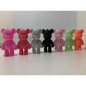 May include: A row of seven bear-shaped figurines covered in small, reflective rhinestones. The bears are arranged in a line and feature a variety of vibrant colours including bright pink, orange-red, silver, black, light pink, light green, and orange.