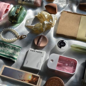 May include: A collection of beauty and stationery products. Items include a pink pouch, a green and gold compact, a pearl bracelet, a green floral headband, a notebook, and face paper. Other items include a makeup sponge, a jade roller, and a small spray bottle.