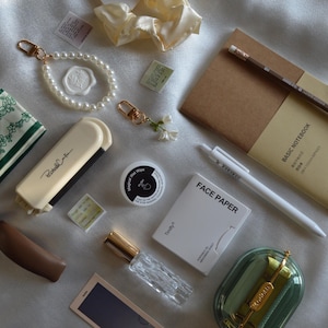 May include: A collection of stationery and accessories, including a pearl bracelet, a notebook, a pen, and a pencil. Other items include a small perfume bottle, a compact mirror, and a small green and white floral patterned handkerchief.