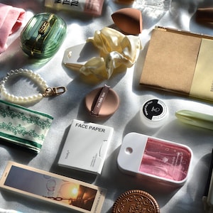 Könnte beinhalten: Eine Auswahl an Schönheits- und Lifestyle-Artikeln, darunter ein rosa Handtuch, ein grüner und goldener Kompaktpuder, ein Perlenarmband, ein grünes Blumenstirnband, ein Notizbuch und Gesichtspapier.