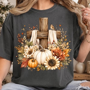 May include: A charcoal gray t-shirt featuring a graphic print of a wooden cross draped with white fabric, surrounded by a cluster of autumn-colored sunflowers, orange pumpkins, fall foliage, and a small bird perched on the cross.