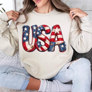 May include: A cream-coloured sweatshirt displaying the word "USA" in a patriotic design. The letters are decorated with the American flag's stars and stripes, with a stitched patchwork effect. The sweatshirt is a casual, comfortable style.