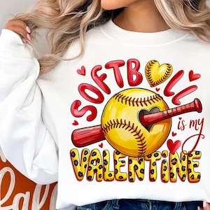 Pode incluir: Camisola branca com um gráfico de softball. O design apresenta uma bola de softball amarela com um recorte em forma de coração, um bastão vermelho e as palavras "Softball is my Valentine" em vermelho e estampa de leopardo.