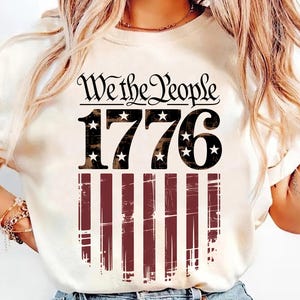 Może przedstawiać: Kremowa koszulka z napisem "We the People" nad numerem "1776" z akcentami gwiazd. Poniżej znajduje się postarzany wzór amerykańskiej flagi w czerwono-białe paski. Koszulka o tematyce patriotycznej.