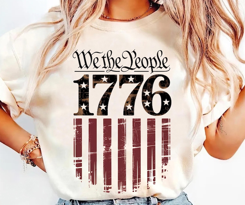 Op de afbeelding: Cr&egrave;mekleurig T-shirt met de tekst "We the People" boven het nummer "1776" met steraccenten. Daaronder een distressed Amerikaans vlagontwerp in rode en witte strepen. Een patriottisch thema T-shirt.