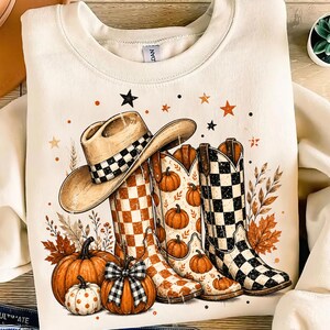 Puede incluir: Una sudadera de cuello redondo color crema con un estampado gráfico de un sombrero de vaquero beige, dos botas vaqueras con patrones de cuadros naranjas y negros, y varias calabazas naranjas, hojas de otoño, espigas y estrellas.