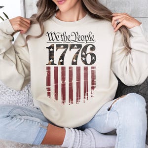 Op de afbeelding: Cr&egrave;mekleurige sweatshirt met de tekst "We the People 1776" boven een versleten Amerikaans vlagontwerp. Het ontwerp heeft rode en witte strepen en sterren in de cijfers. Een casual, patriottische stijl.