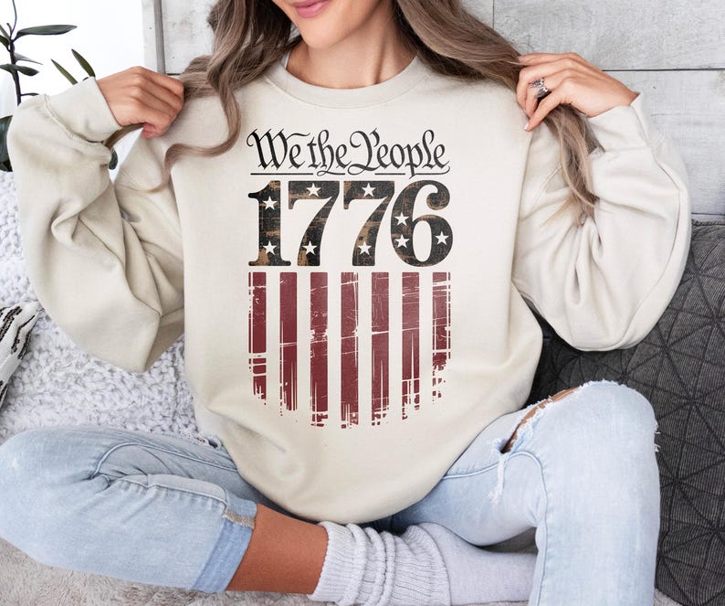 Op de afbeelding: Cr&egrave;mekleurige sweatshirt met de tekst "We the People 1776" boven een versleten Amerikaans vlagontwerp. Het ontwerp heeft rode en witte strepen en sterren in de cijfers. Een casual, patriottische stijl.