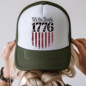 Op de afbeelding: Een truckerpet met een witte voorkant en een donkergroene rand en mesh achterkant. De voorkant heeft de tekst "We the People 1776" boven een versleten Amerikaans vlagontwerp.