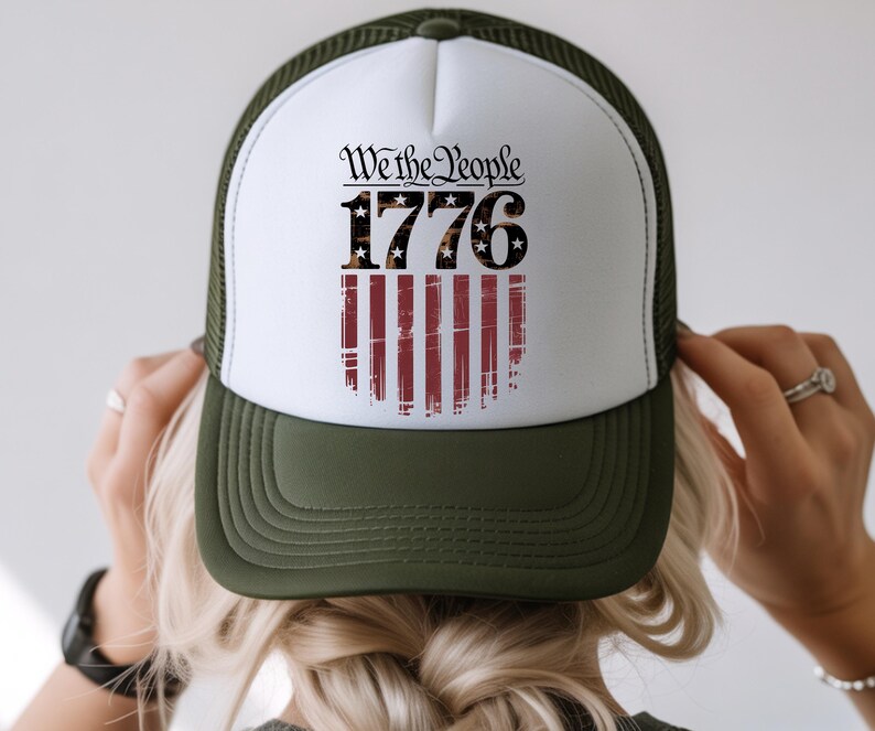 Op de afbeelding: Een truckerpet met een witte voorkant en een donkergroene rand en mesh achterkant. De voorkant heeft de tekst "We the People 1776" boven een versleten Amerikaans vlagontwerp.