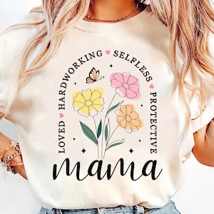 May include: Cream-colored t-shirt featuring a floral design with the word "mama" in a script font. The design includes yellow, pink, and orange flowers, a butterfly, and the words "loved, hardworking, selfless, protective".