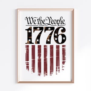 Op de afbeelding: Ingelijste print met de tekst "We the People" boven het jaar "1776" en een versleten Amerikaans vlagontwerp met rode en witte strepen. De print zit in een goudkleurige lijst.