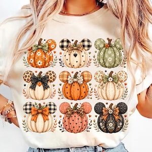 Op de afbeelding: Een crèmekleurig t-shirt met een raster van negen geïllustreerde pompoenen, elk versierd met muizenoorvormen, strikken en diverse patronen zoals bloemen, ruiten, luipaardprint en stippen in tinten oranje, groen, zwart en wit.