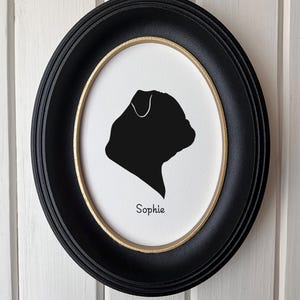 May include: An oval wall frame with a black outer border and a thin gold inner rim. Inside, a white background features a black silhouette of a dog head in profile, with the name Sophie printed in black text underneath the silhouette.