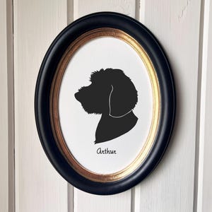 May include: An oval framed silhouette of a dog's head in black, with the name "Arthur" in script below. The frame is black with a gold inner edge, mounted on a white wooden background.