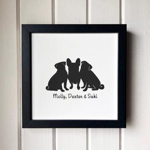 May include: A square black frame containing a white print with the black silhouettes of three dogs sitting side by side. Below the dogs, the text Molly, Dexter & Suki is printed in a simple black font. The artwork is centered within the frame.