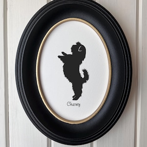 May include: An oval wall frame with a black outer border and a thin gold inner rim. Inside, a white background features a black silhouette of a small, fluffy dog standing on its hind legs, with the name Chewy printed in black text below.