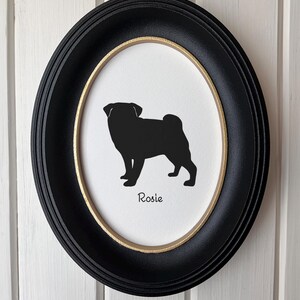May include: An oval wall frame with a black outer border and a thin gold inner rim. Inside, a white background features a black silhouette of a pug dog standing in profile, with the name Rosie printed in black script lettering below the dog.