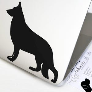 May include: Black silhouette of a German Shepherd dog on a white background. The silhouette is facing left and has its tail curled around its legs. The image is on a laptop with a business card for Silhouette My Pet.