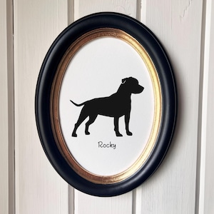 May include: An oval framed print featuring a black silhouette of a dog on a white background. The frame is black with a gold inner edge. The dog's name, "Rocky," is printed in black below the dog.