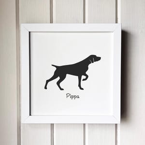 May include: A framed print featuring a black silhouette of a dog in a walking pose. The dog's name, "Pippa," is written in a script font below the image. The print is set in a white square frame against a white wooden background.