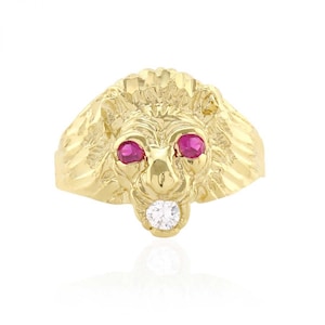May include: A gold-colored lion ring with ruby eyes and a diamond in its mouth. The ring is a detailed sculpture of a lion's head, showcasing a textured mane and facial features. The ring is set against a white background.