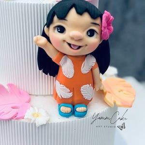 May include: A cake decoration featuring a smiling cartoon girl in an orange dress with white feather accents and blue sandals. She has black hair and a pink flower. The cake is white with pink and yellow floral accents. The text "Yumm Cake ART STUDIO" is visible.