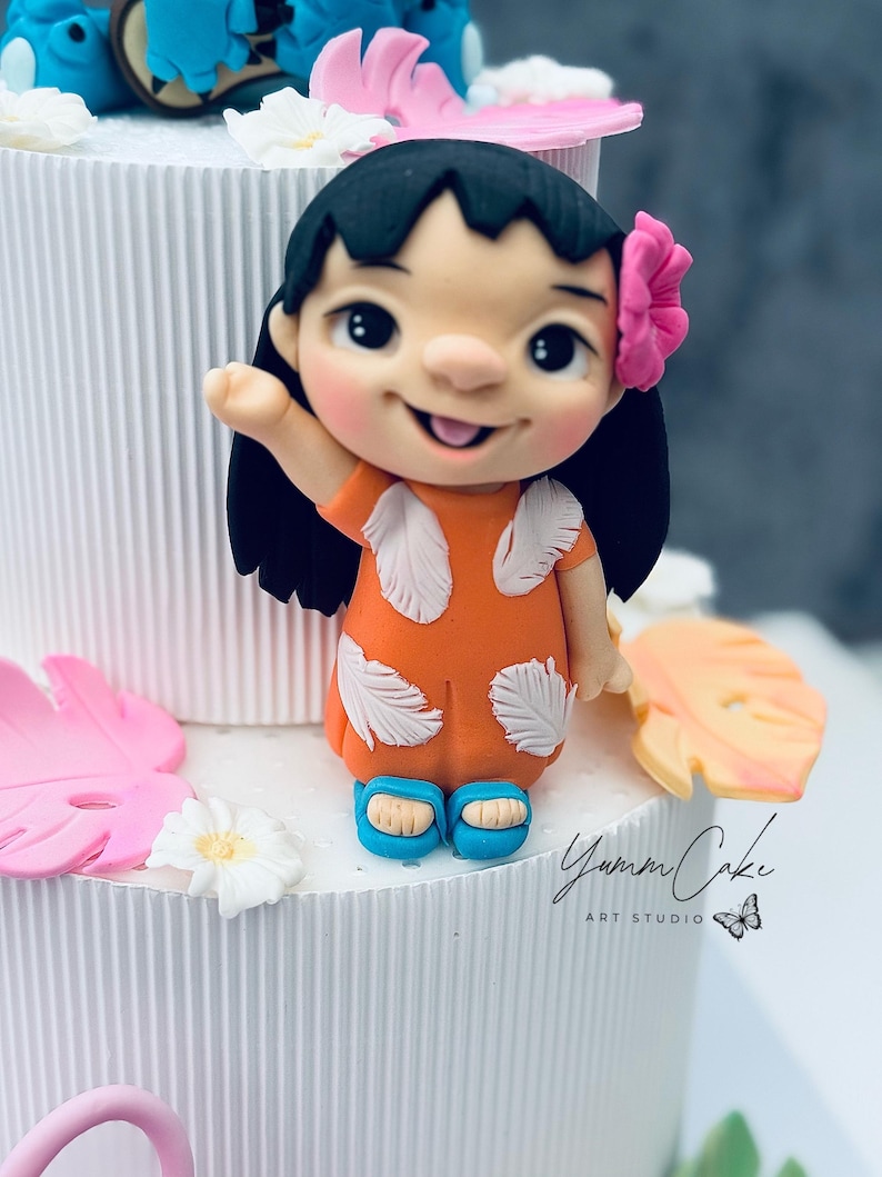May include: A cake decoration featuring a smiling cartoon girl in an orange dress with white feather accents and blue sandals. She has black hair and a pink flower. The cake is white with pink and yellow floral accents. The text "Yumm Cake ART STUDIO" is visible.