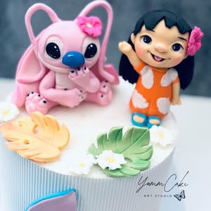 May include: A decorated cake featuring two cartoon characters, one pink with a flower and the other in an orange dress. The cake is white with blue vertical stripes and decorated with leaves and flowers. The text "Yumm Cake Art Studio" is visible.