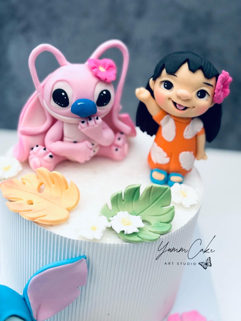 May include: A decorated cake featuring two cartoon characters, one pink with a flower and the other in an orange dress. The cake is white with blue vertical stripes and decorated with leaves and flowers. The text "Yumm Cake Art Studio" is visible.