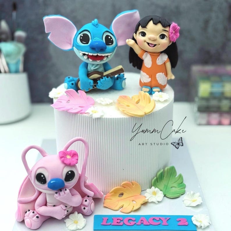 May include: A white cake decorated with figures of Stitch, Lilo, and Angel. Stitch is blue and playing a ukulele. Lilo is wearing an orange dress. Angel is pink. The cake is decorated with flowers and leaves. The text "Legacy 2" is on a blue banner.