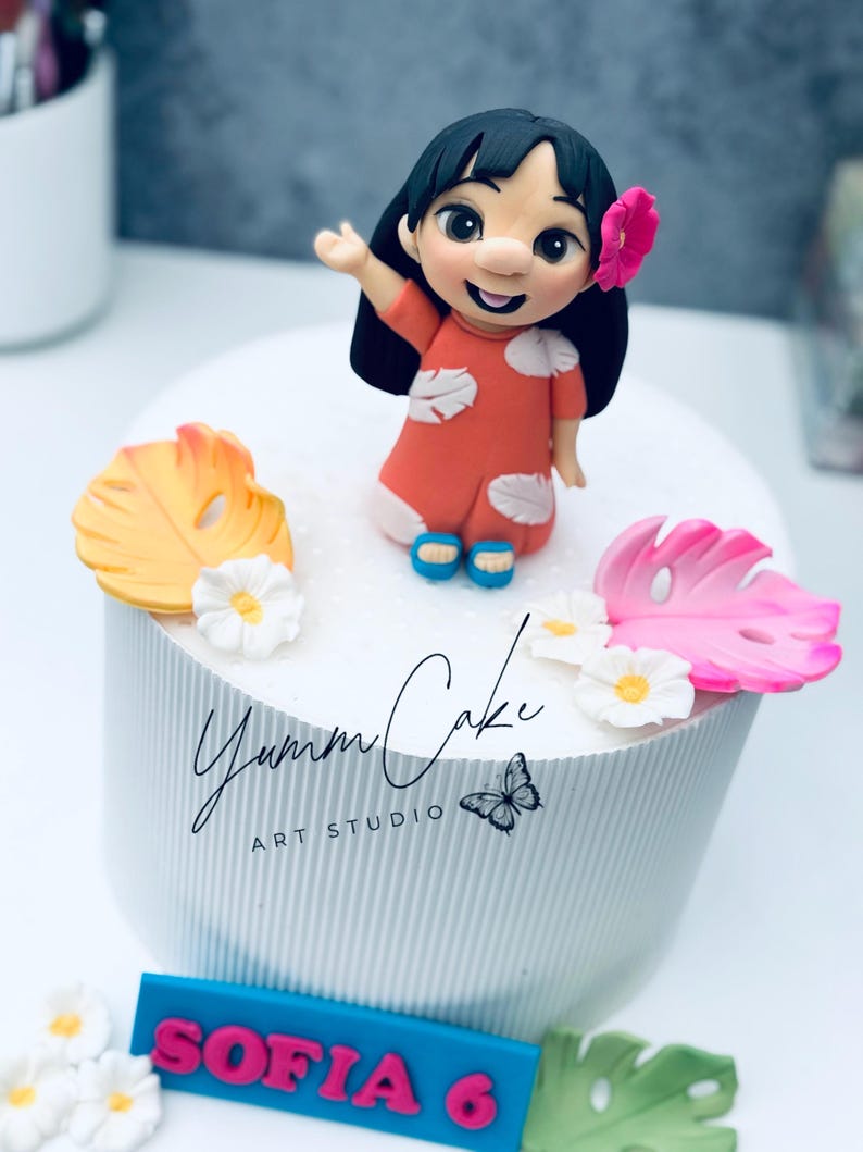 May include: A custom-made cake featuring a figurine of a cartoon character in an orange dress, with a pink flower in her hair. The cake is decorated with tropical leaves and flowers, and the name "Sofia 6" is displayed on a blue plaque.