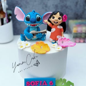 May include: A white cake with a blue and pink nameplate that reads "SOFIA 6". The cake is decorated with figurines of Stitch playing a ukulele and Lilo, along with floral and leaf accents. The cake has the text "Yumm Cake ART STUDIO".